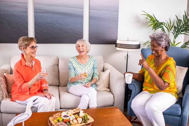 Three elderly women sit together in a cozy living room, enjoying drinks and laughing. A coffee table in front of them has a platter of snacks. Bright artwork and a plant decorate the room.