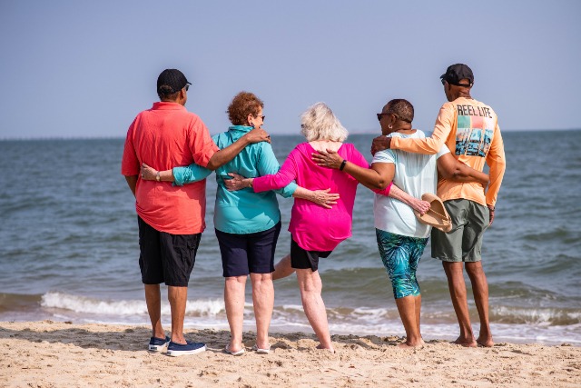 Five people stand arm in arm, facing the ocean on a sandy beach. They wear casual, colorful clothing, and it is a sunny day. The mood appears relaxed and joyful.