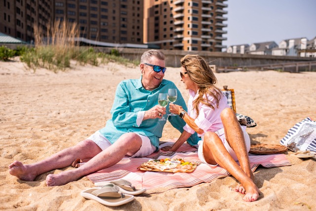 A man and woman sit on a beach blanket, toasting with wine glasses. They are smiling and enjoying a sunny day. Sandals and a picnic tray with snacks are visible. Sandy beach and buildings are in the background.