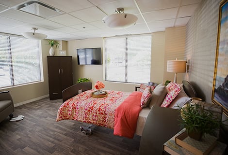 A hospital room featuring a bed with floral bedding, a TV mounted on the wall, a dark wood cabinet, and a window with blinds. There are plants, a chair, and a lamp, creating a comforting and welcoming atmosphere.