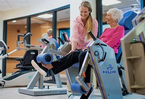 An elderly woman in a pink sweater uses a leg exercise machine, assisted by a smiling female fitness trainer in a light pink shirt. They are in a gym setting with various exercise equipment in the background.