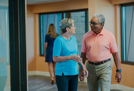 An older man and woman are walking and talking indoors, both wearing casual clothes. The man wears a pink shirt and glasses, and the woman is in a blue shirt. A person in a dress is walking away in the background.