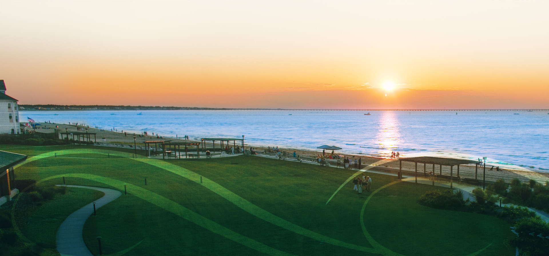 A scenic view of a beach at sunset with a vibrant orange sky. The sun is setting over the horizon, casting reflections on the calm sea. A grassy area with pathways and wooden shelters is in the foreground, with people walking along the shoreline.