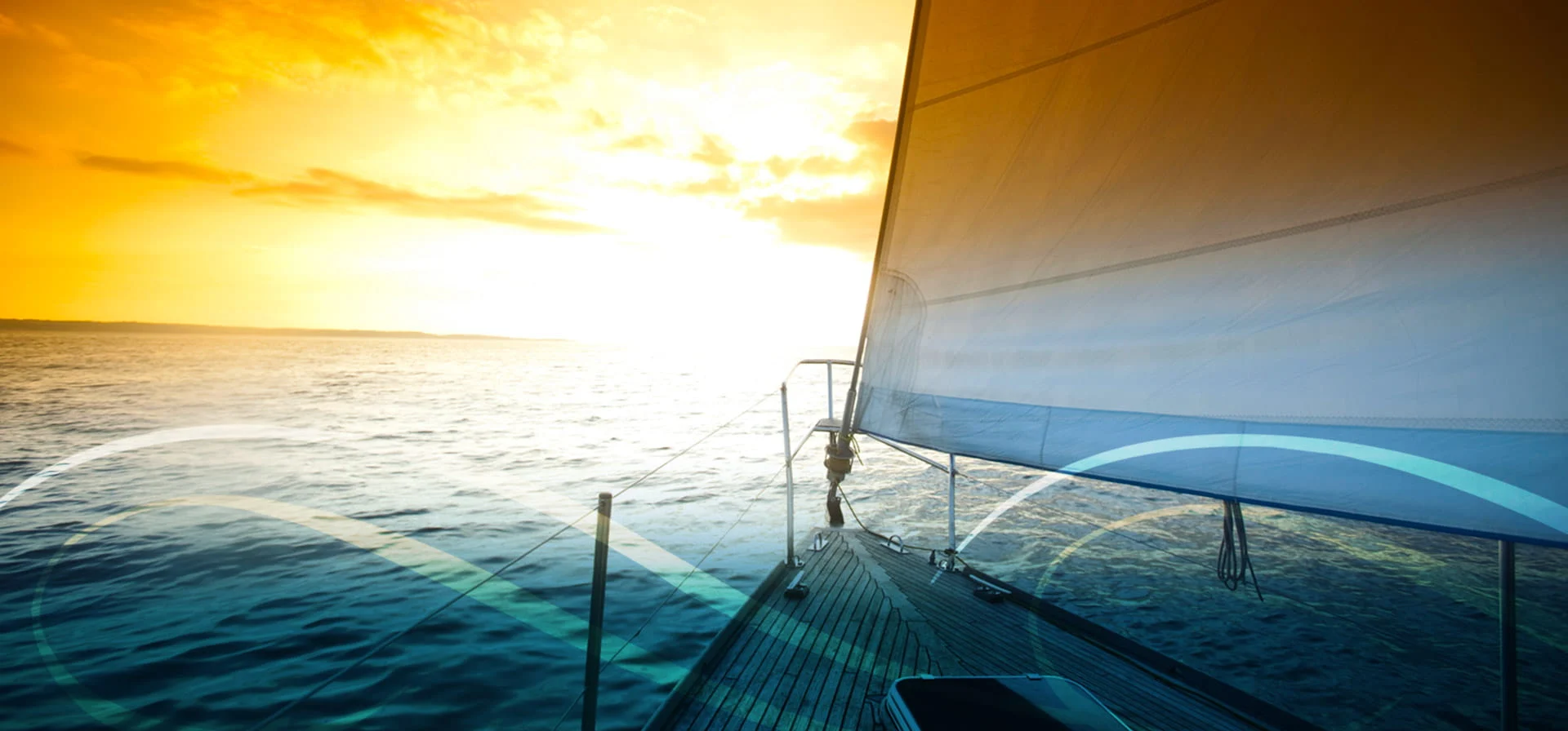 A sailboat gliding on the ocean at sunset, with warm orange and yellow hues illuminating the sky. The sails are partly visible, casting a shadow on the deck. Calm waters extend to the horizon.