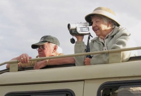Two older adults are standing on an open-top vehicle. The woman is smiling and using a video camera, while the man looks outward. Both are wearing hats and casual outdoor clothing, suggesting they are on a safari or tour.