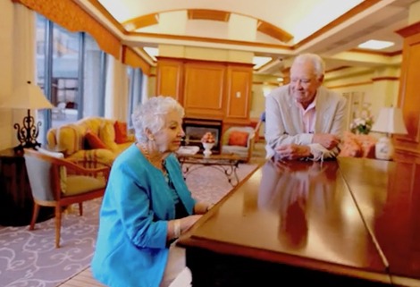 An elderly woman wearing a blue jacket plays the piano while an elderly man in a suit watches in a cozy, well-lit room with comfortable seating and elegant decor.