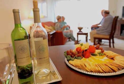 A table with two bottles of wine and a platter of crackers, cheese, and grapes. In the background, four people sit on a sofa, engaged in conversation in a cozy living room.