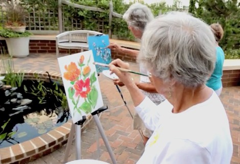 An elderly person with gray hair paints colorful flowers on a canvas using a brush, seated by a pond and garden. Another person stands nearby, painting on a different canvas.