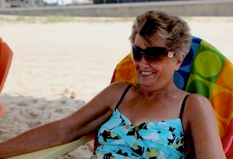 A smiling woman with short hair wearing sunglasses and a floral swimsuit sits in a colorful beach chair on a sandy beach.