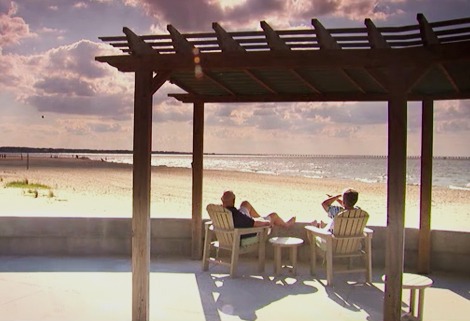 Two people relax on wooden chairs under a pergola by a sandy beach. They are facing the ocean, enjoying the sunny day with a partly cloudy sky. A long pier is visible in the distance.