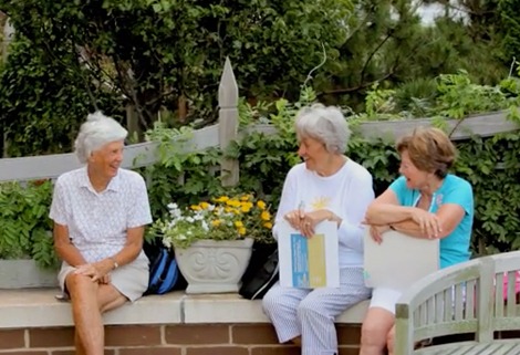 Three elderly women are sitting on a bench outdoors, engaged in conversation. They are surrounded by greenery and have a planter with yellow and white flowers in front of them. One woman holds a book, while another has a folder. They appear relaxed and happy.