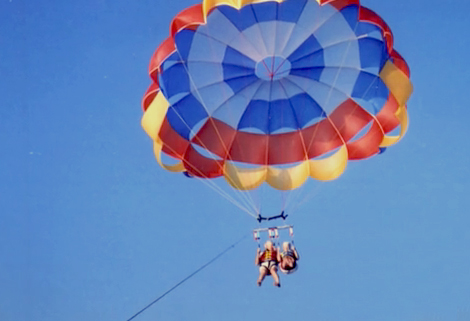 Two people parasailing with a colorful parachute featuring blue, red, and yellow sections against a clear blue sky. They are suspended in harnesses, high above, connected to a boat by a long cable.