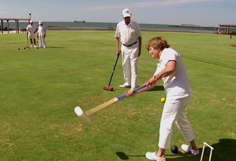 A group of people dressed in white play croquet on a grassy field near the ocean. One person is swinging a mallet, while others watch. The scene is sunny with a blue sky and distant piers in the background.