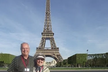 An elderly couple stands smiling in front of the Eiffel Tower on a clear day.