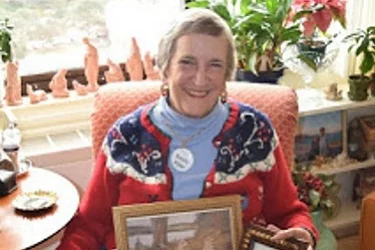 A smiling elderly woman sits in a cozy room, holding two framed photos. Shes wearing a red sweater with a patterned shawl and a round name badge. The background is filled with plants and decorative items on a windowsill.