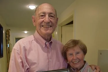 An elderly couple smiles warmly in a well-lit hallway. The man holds a picture frame while they both wear pink shirts. The background features open doors and soft lighting.