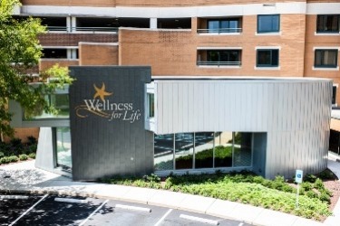 Modern wellness center with a sign reading Wellness for Life on its exterior. The building features a mix of brick and metallic finishes with large windows. A tree and greenery are visible in the foreground, adding a touch of nature.