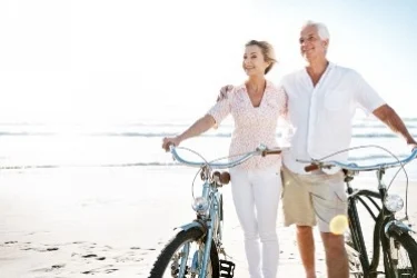 An older couple walks their bicycles on a sunny beach, smiling and enjoying the day. The ocean is visible in the background under a clear sky.