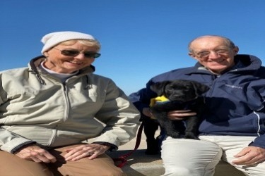 An elderly couple, both wearing sunglasses, sits outdoors on a sunny day. The woman wears a beanie and jacket, while the man wears a cap and jacket. They are holding a small black puppy. The sky is clear and blue.
