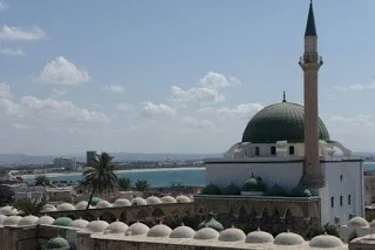 A mosque with a green dome and tall minaret stands against a backdrop of a coastal cityscape under a blue sky with scattered clouds.