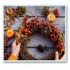 A person arranges a festive wreath made of twigs, red berries, and orange autumn leaves on a wooden surface. Two small pumpkins and a spool of black thread are nearby.