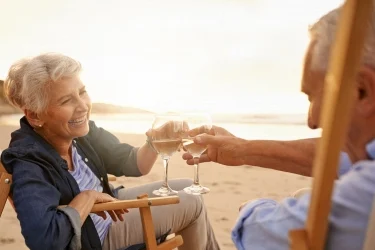An elderly couple sitting in wooden chairs on a beach clink wine glasses, smiling at each other. The sun is setting in the background, casting a warm glow over the scene.