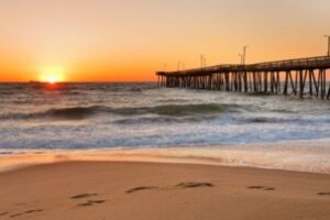 Sunset over a beach with gentle waves and a sandy shore. A pier extends into the ocean from the right, and the sun casts a warm, orange glow across the sky and water. Footprints are visible in the sand in the foreground.