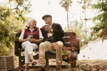 An elderly couple sits on a park bench holding coffee cups, enjoying a fall day. Leaves are falling around them, and they are both smiling. The scene is surrounded by trees with autumn foliage, creating a warm and relaxed atmosphere.