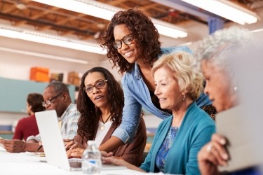 A group of adults in a classroom setting, with a woman standing and assisting others who are seated. They are focused on a laptop and appear engaged in a learning activity.