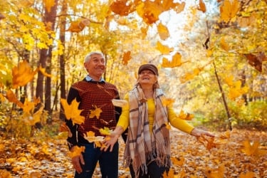 An elderly couple joyfully tosses autumn leaves in the air while walking through a forest. They are surrounded by vibrant yellow and orange foliage, creating a warm, cheerful atmosphere.