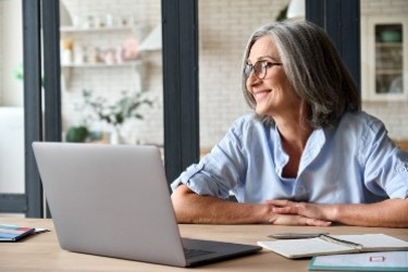 A woman with gray hair and glasses sits at a table in front of a laptop, smiling. She is wearing a light blue shirt. In the background, there are shelves with plants and books in a cozy room.