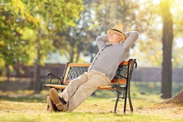 An elderly man in a hat and sweater relaxes on a park bench with his hands behind his head. The sun shines through the trees, creating a peaceful atmosphere in a grassy, tree-filled park.