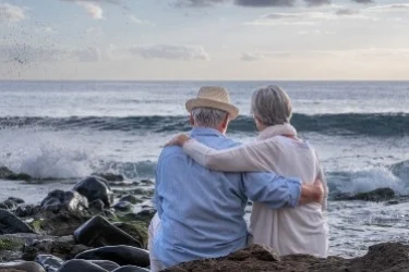 A senior couple sits on rocks by the ocean, embracing and gazing at the waves under a cloudy sky. The man wears a straw hat and blue shirt, while the woman in a light sweater rests her head on his shoulder.