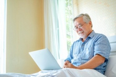An older man with gray hair and glasses sits in bed, smiling while using a laptop. He is wearing a blue shirt, and sunlight filters through a window, creating a warm and relaxed atmosphere.