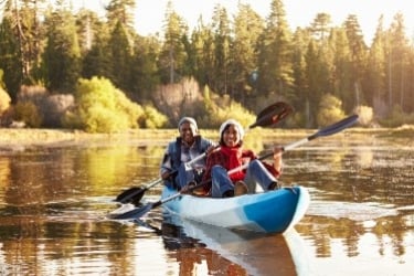 Two people paddle a blue kayak on a calm lake surrounded by a forest. They are wearing casual outdoor clothing and life jackets, enjoying a sunny day on the water with trees reflecting in the lake.