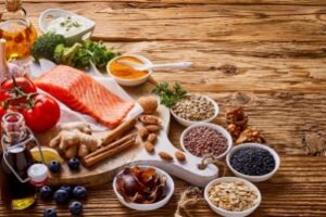 A wooden table displays ingredients including salmon, broccoli, tomatoes, grains, spices, nuts, blueberries, and oil. Assorted seeds and spices are in small dishes, showcasing a variety of healthy foods.