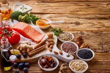 A wooden table displays ingredients including salmon, broccoli, tomatoes, grains, spices, nuts, blueberries, and oil. Assorted seeds and spices are in small dishes, showcasing a variety of healthy foods.