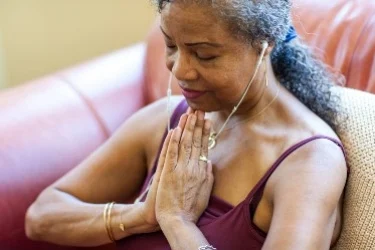 A woman sitting on a couch, eyes closed, wearing earphones, and with her hands in a prayer position. Her expression is calm and relaxed, suggesting meditation or deep concentration. She is wearing a sleeveless top.
