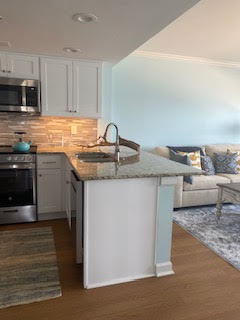 Modern kitchen with white cabinets, granite countertops, and stainless steel appliances. A cozy living area with a beige sofa and patterned cushions is visible in the background, along with a blue accent wall.