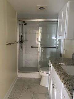 A modern bathroom with a walk-in shower featuring glass doors and tiled walls. Next to the shower is a toilet, and a granite countertop with a sink on the right. The floor is covered in light-colored tiles.