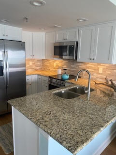 A modern kitchen with granite countertops, white cabinets, and a stainless steel refrigerator. The backsplash is tiled in a light brown and beige pattern. A double sink is installed in the island with a polished faucet, and a kettle is on the stove.