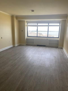 An empty room with light-colored walls and a large window. The floor has a wood-like finish. The window offers a view of the ocean, with some buildings visible in the distance.