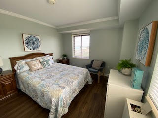 A cozy bedroom with a double bed featuring floral bedding, two nightstands, a window with blinds, a blue armchair, and decorative art on the walls. A dresser with books and a potted plant completes the tranquil setting.