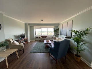 A bright living room with large windows showing an outside view. The room features wooden flooring, a green rug, and seating including armchairs and a sofa. Plants and a table with decorations complete the cozy, modern design.