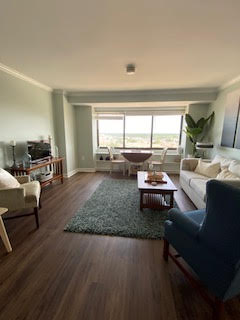 A cozy, well-lit living room with wooden floors features a cream sofa, blue armchair, and a central wooden coffee table on a green rug. A large window shows a scenic view, while a TV and plants add to the decor.