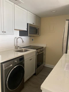 A modern kitchen with white cabinets, a stainless steel oven, a microwave, and a refrigerator. Theres a sink with a faucet and a washing machine under the counter. The kitchen has light-colored walls and a white countertop.
