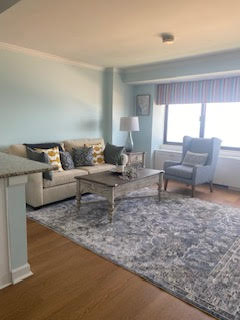 A cozy living room with a light blue wall, beige sofa with accent pillows, gray armchair, and wooden coffee table on a patterned rug. Theres a window with a striped valance, and a table lamp adds a warm touch to the space.