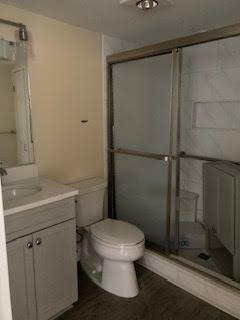 A small bathroom with beige walls features a gray vanity with a sink and mirror, a white toilet, and a shower with frosted glass sliding doors. The shower has white tile walls and a built-in shelf. The floor is covered with dark laminate.