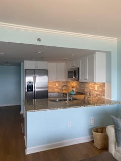 A modern kitchen with light blue walls and white cabinets. It features a granite countertop, stainless steel appliances, including a refrigerator and microwave, and a tile backsplash. A woven basket is on the floor next to a gray couch.