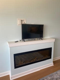 A flat-screen TV is mounted on a light blue wall above a white mantel with a built-in electric fireplace. The room has hardwood floors and a light-colored area rug in front of the fireplace.